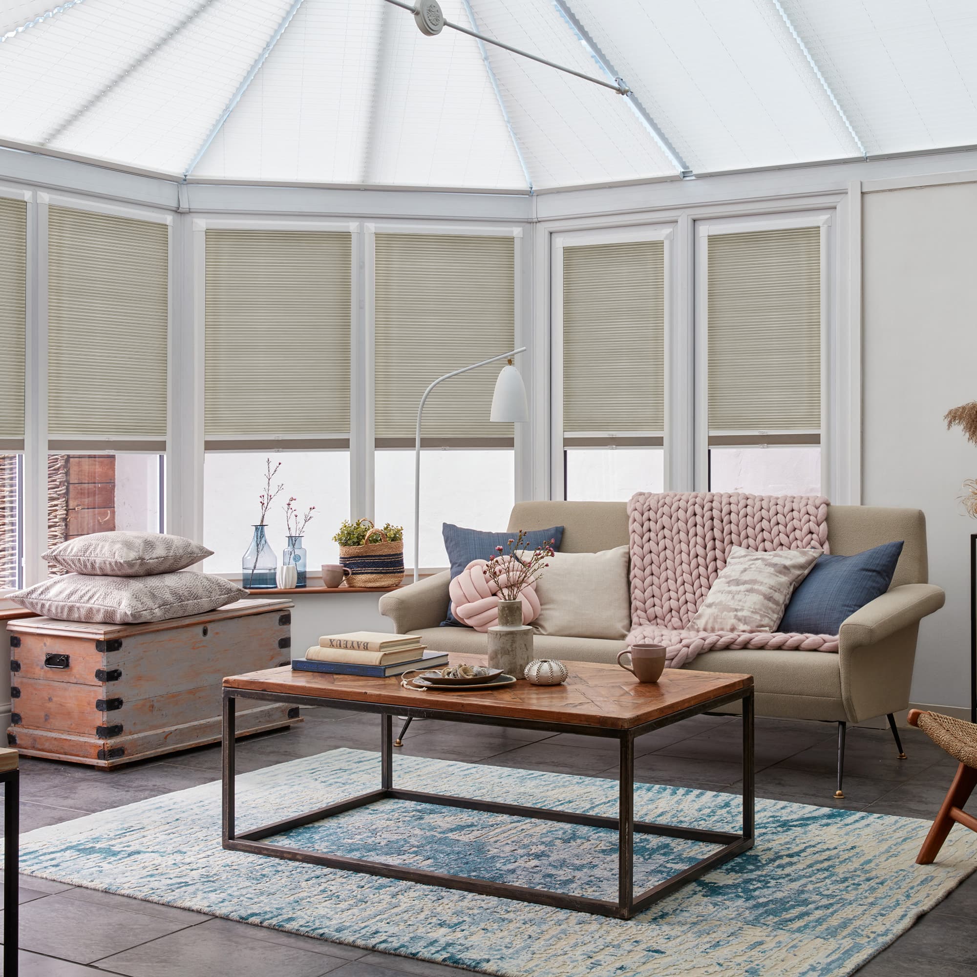 Beige pleated cellular blinds covering tall conservatory windows, mostly lowered and filtering soft daylight; a cozy sunroom with a sofa, knitted throw, wooden coffee table and pale blue rug.