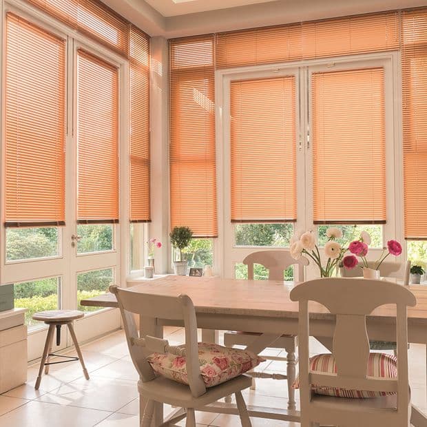 Peach-colored horizontal Venetian blinds cover tall glass doors and windows, mostly lowered and filtering warm daylight; dining nook with a wooden table, chairs with floral cushions, vases, and garden view.