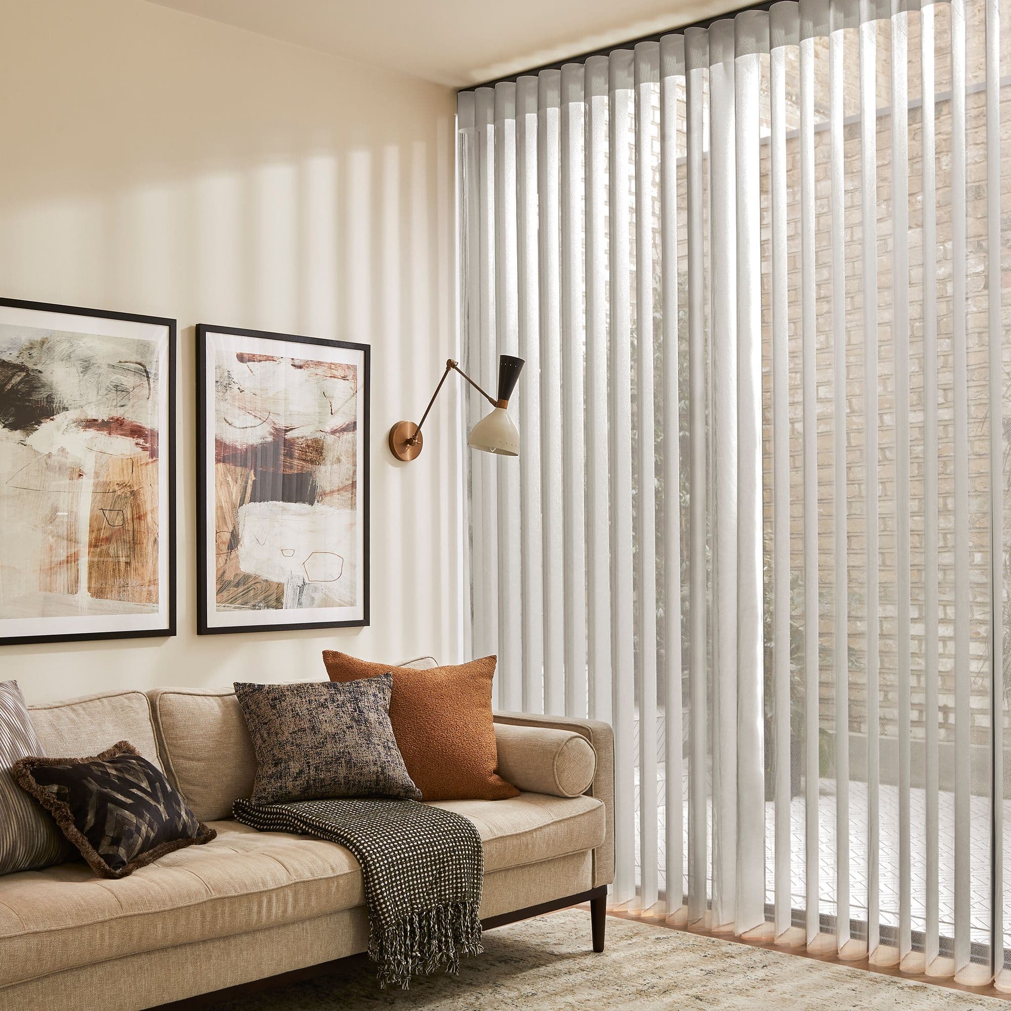 Vertical sheer fabric blinds span floor-to-ceiling sliding glass doors, gently filtering daylight; modern living room with beige sofa, textured cushions, abstract wall art, brass sconce, and brick courtyard outside.