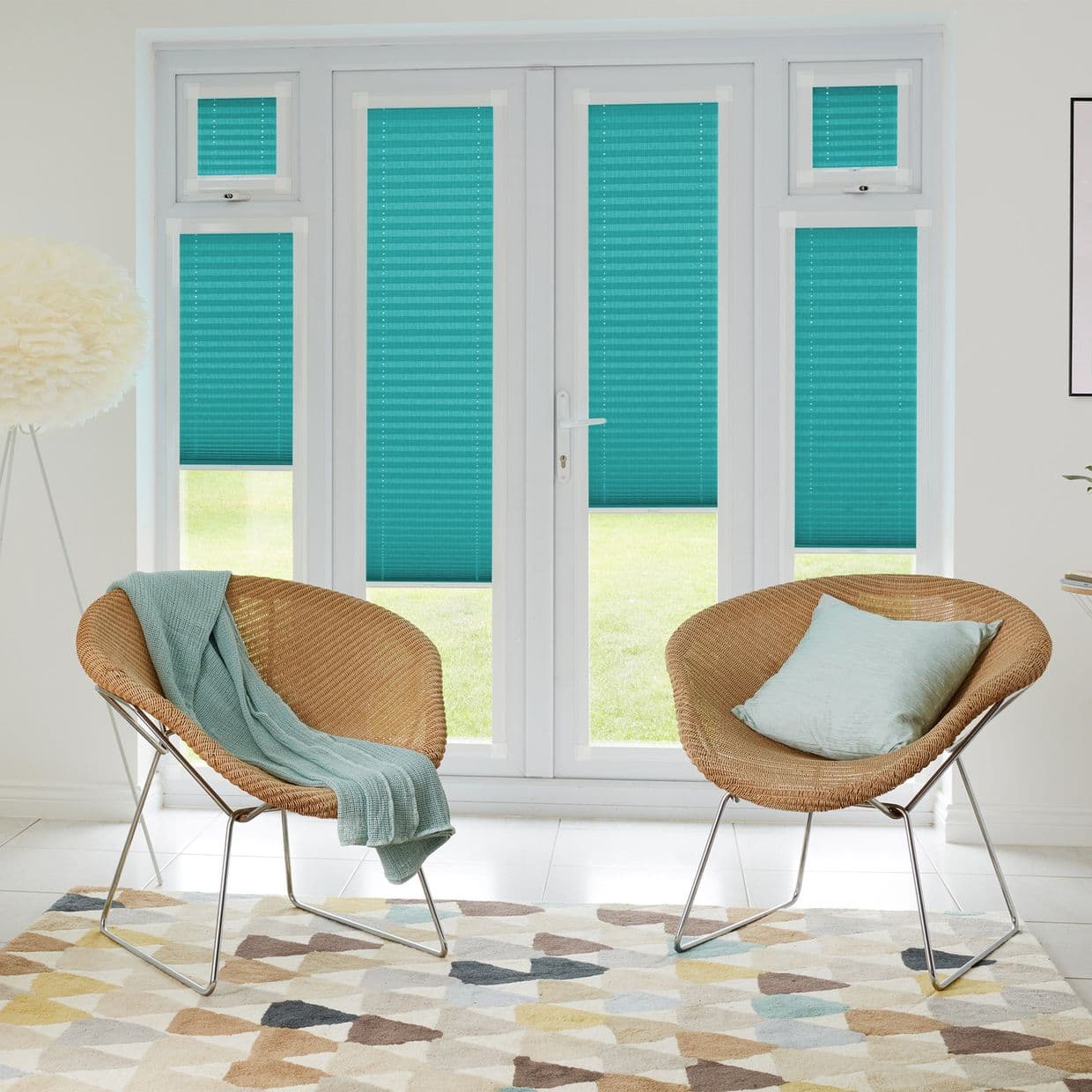 Turquoise pleated blinds cover a white-framed French door and four surrounding windows, mostly lowered and filtering daylight. Bright sitting room with two wicker chairs, patterned rug, and garden view.