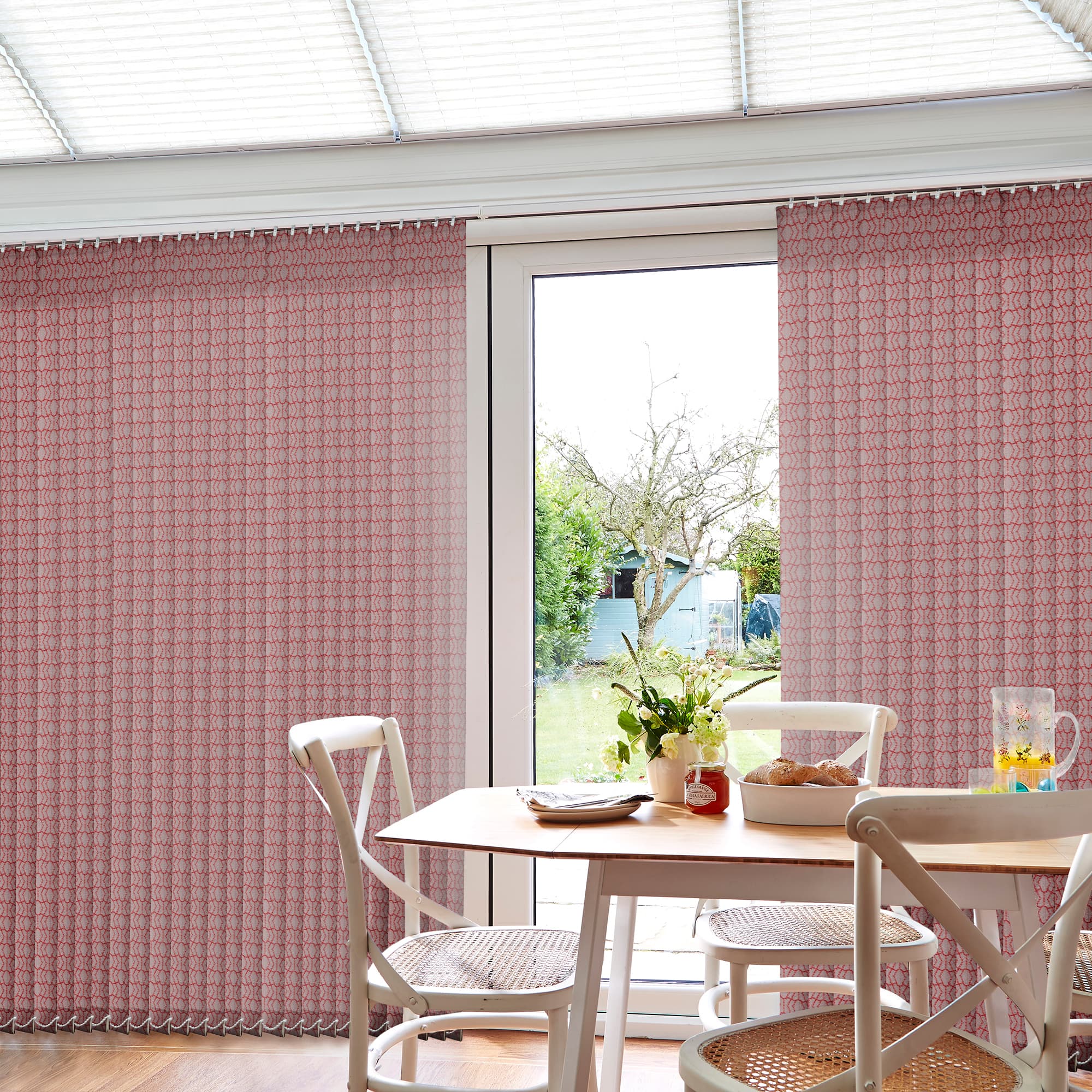 Patterned pink vertical blinds span a patio door, partially drawn to either side, filtering daylight; dining table with chairs, flowers, bread and jug sits inside, bright garden visible outside.