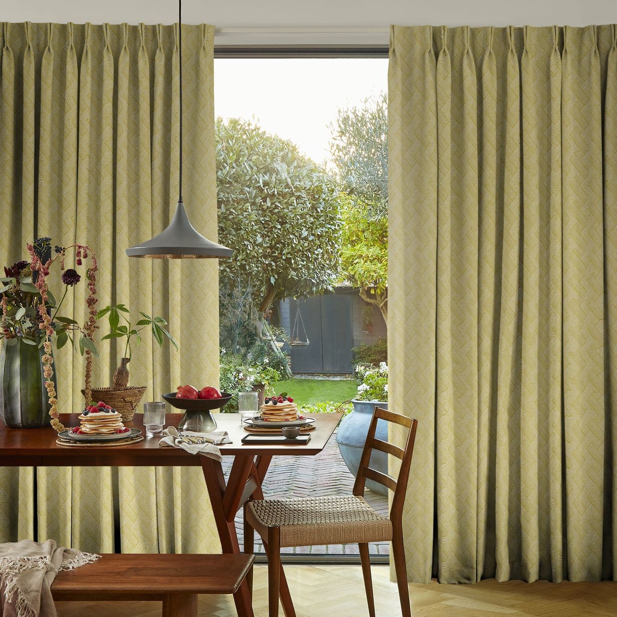 Pleated pale-green patterned curtains, hung on either side of a sliding door, are parted to reveal daylight; a breakfast-set dining table sits inside, opening onto a sunlit garden.