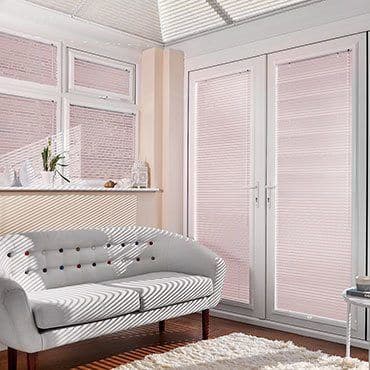 Pleated pale-pink blinds cover bay windows and French doors, fully lowered and filtering daylight, casting striped shadows across a white tufted sofa and shag rug in a sunlit living room.