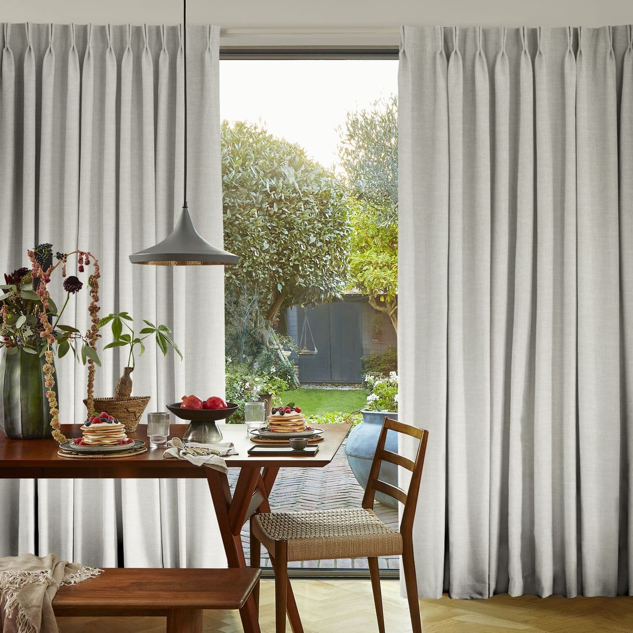 Floor-length light-gray pleated curtains, parted to reveal a sliding door, filtering soft daylight; a wooden dining table with plates and a pendant lamp faces a sunlit garden with trees.