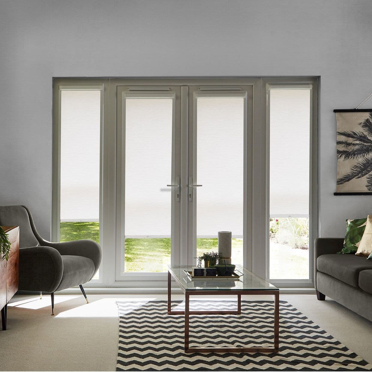 White roller blinds covering double French doors and sidelights, lowered to the sill with a narrow bottom gap, filtering daylight; living room with sofas, glass coffee table and chevron rug.