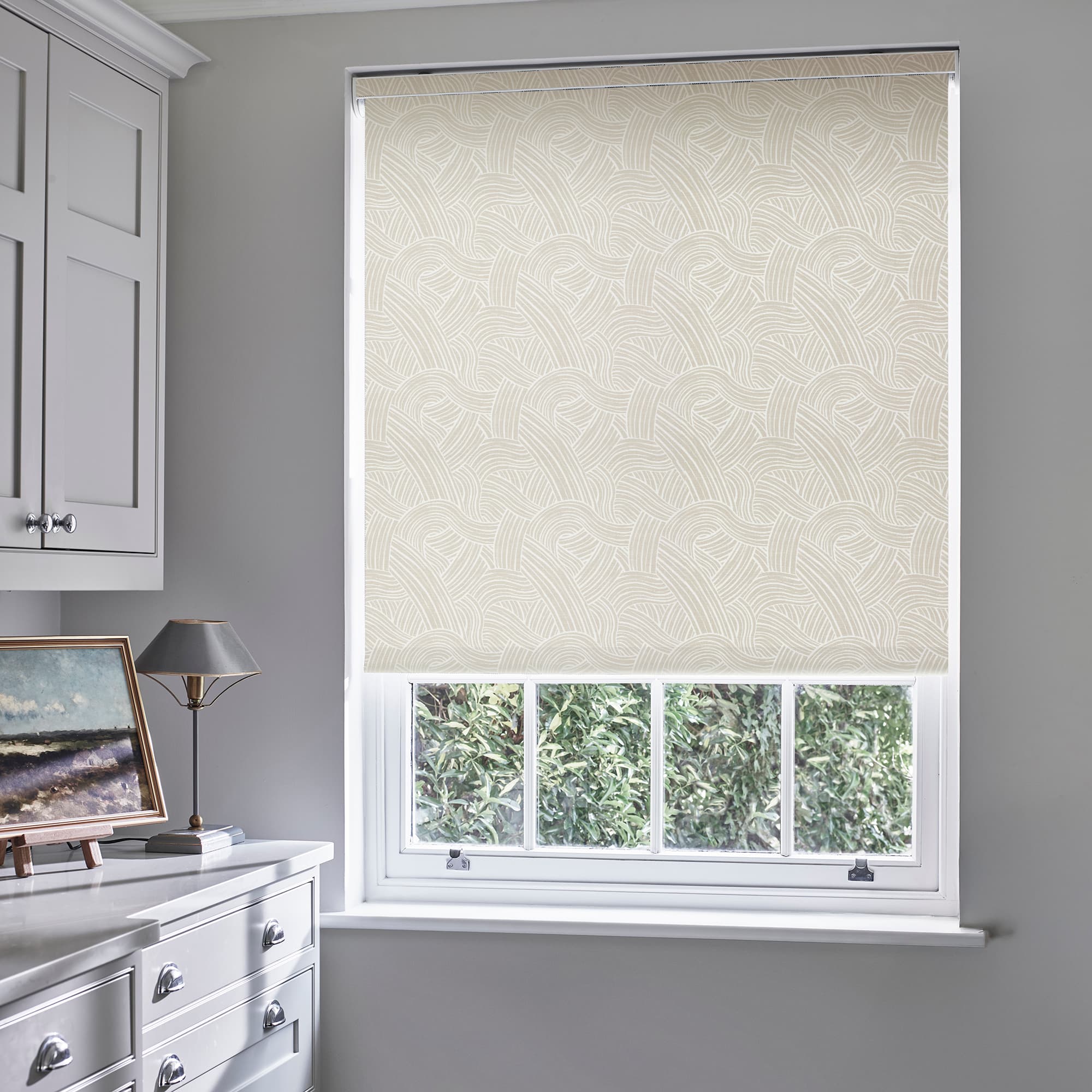 Beige patterned roller blind fully lowered, woven-arc motif filtering daylight; fitted inside a white-trim sash window in a softly lit gray study/dressing area, with cabinets, a lamp and painting beside.