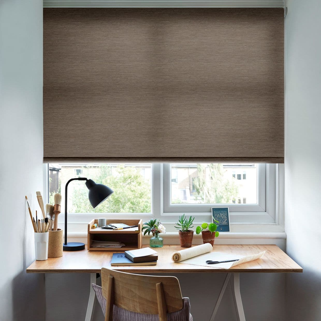 Brown textured roller blind, lowered almost fully across the window, blocking and diffusing daylight; above a wooden desk with a lamp, art supplies, small potted plants and a suburban view.
