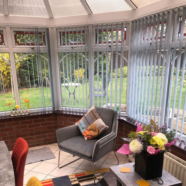 Vertical pale-blue vertical blinds on wraparound conservatory windows, slats partially tilted to filter daylight; a gray armchair with cushion sits on tiled floor in a brick-sided sunroom overlooking a lawn.