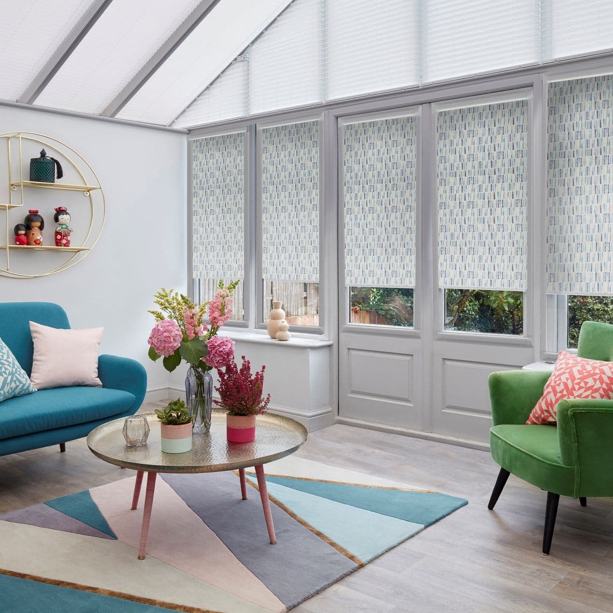 Patterned pastel roller blinds covering tall conservatory doors and upper windows, lowered to filter daylight; in a bright sunroom with teal sofa, green armchair, round coffee table and floral arrangements.