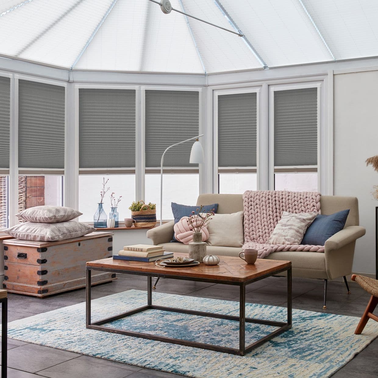Grey pleated cellular blinds, lowered across tall conservatory windows, filtering daylight; beige sofa with pink chunky throw and cushions, wooden coffee table on blue patterned rug in sunlit sitting room.