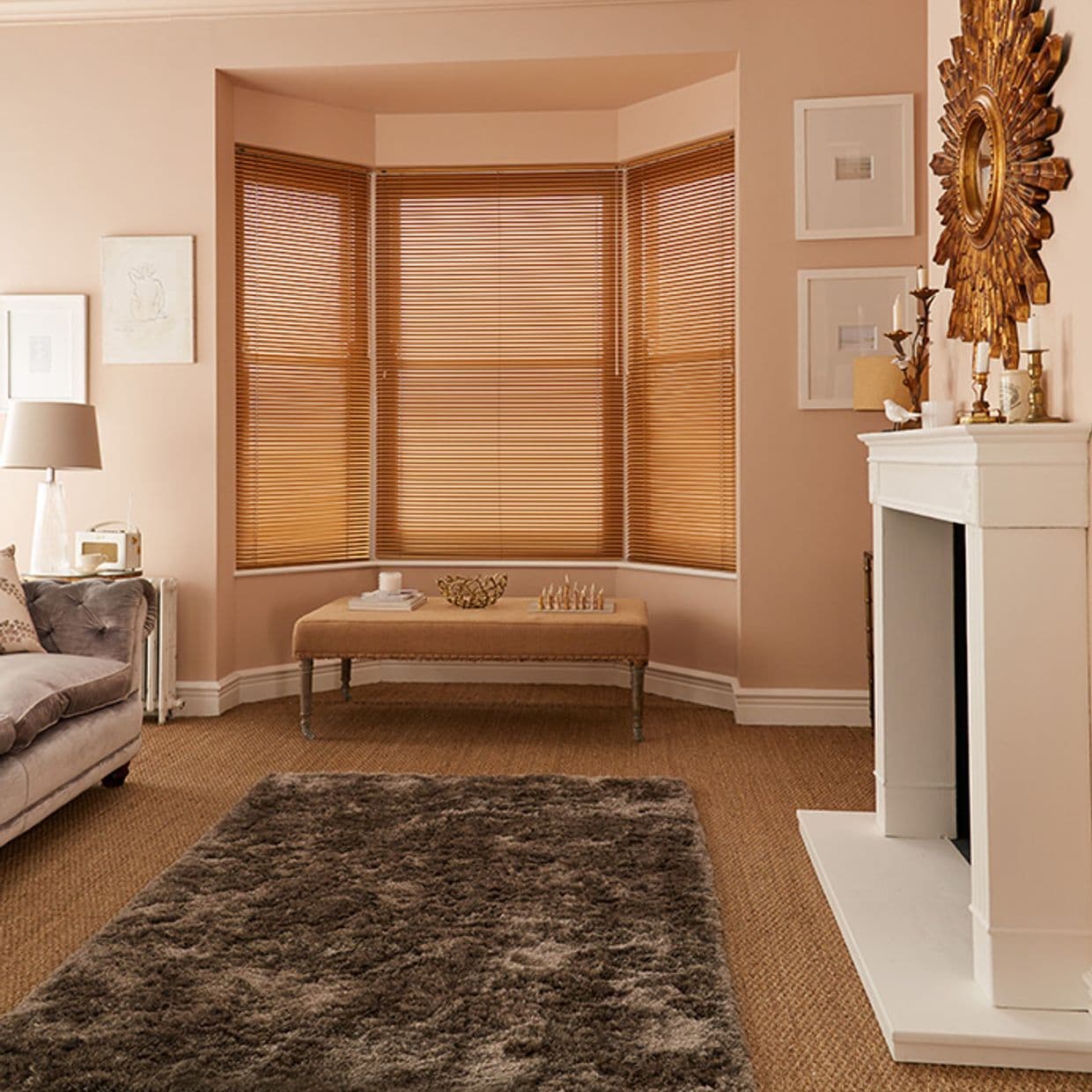 Wooden horizontal blinds on a three-panel bay window, closed and filtering warm daylight; in a softly lit beige living room with tufted sofa, bench, shag rug and white fireplace.