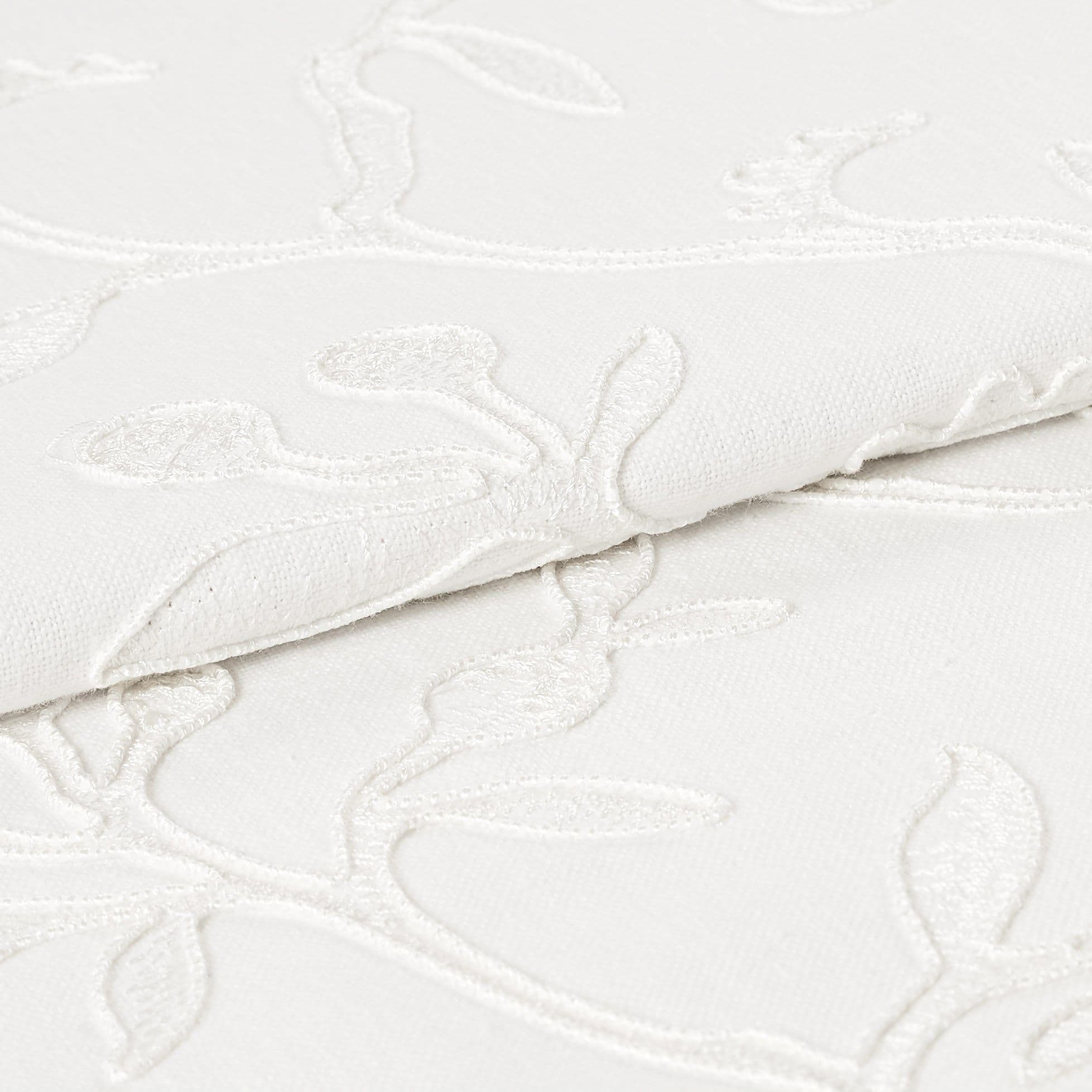 White embroidered fabric folded and overlapping, showing raised floral vine stitching and textured weave; close-up studio view with soft, even lighting emphasizing subtle shadows and fabric detail.