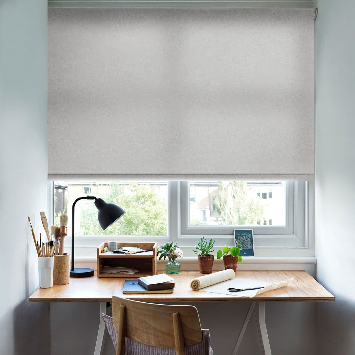 A pale gray roller blind fully lowered across a double window, gently diffusing daylight; a wooden desk beneath holds a lamp, potted plants, brushes and notebooks in a bright home workspace.