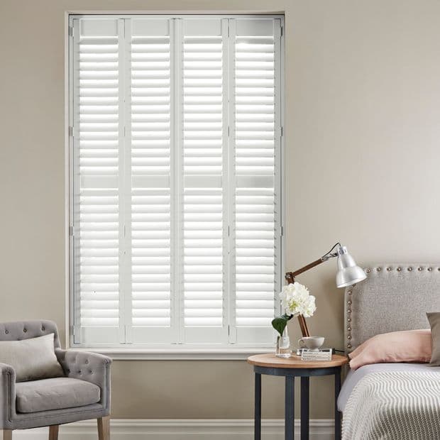 White plantation shuttersâ€”four hinged panels with horizontal louversâ€”cover a tall window; closed, filtering daylight into a neutral bedroom with upholstered bed, bedside table, lamp, armchair, and flowers.