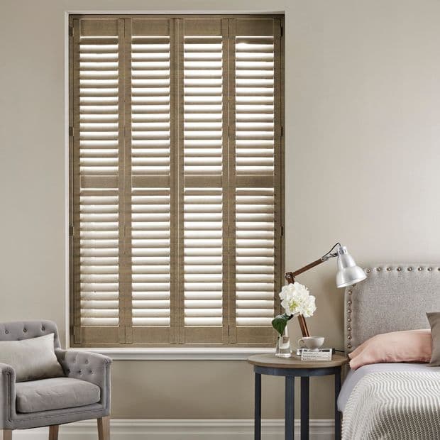 Wooden plantation shutters: four-panel, floor-to-head window covering with horizontal slats. Slats partly angled, filtering soft daylight. Context: neutral bedroom with upholstered bed, side table, lamp, armchair.
