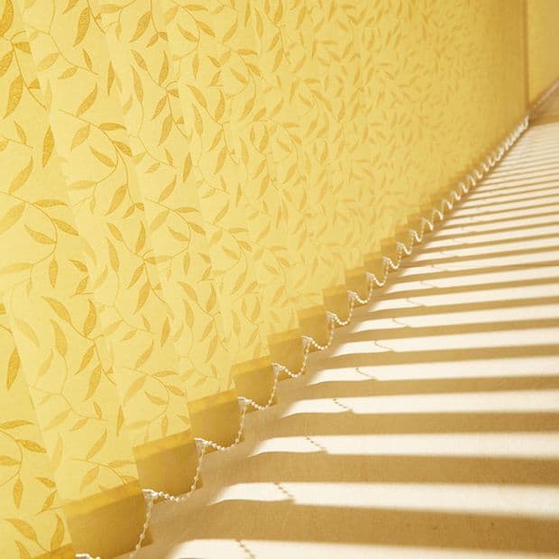 Vertical fabric blinds arranged along a side wall, scalloped bottoms linked by a bead chain, filtering strong sunlight into parallel stripes across a floor beside yellow leafâ€‘pattern wallpaper.
