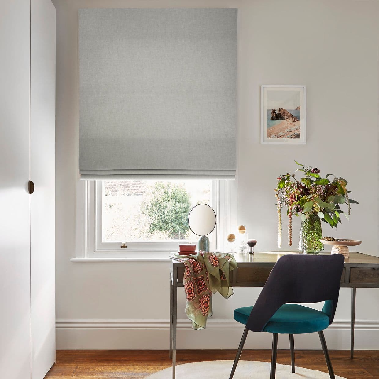 Light-gray fabric Roman blind, lowered above the sill with soft folds, filtering diffuse daylight through a narrow bottom gap; home desk with teal chair, vase of flowers and framed seascape.