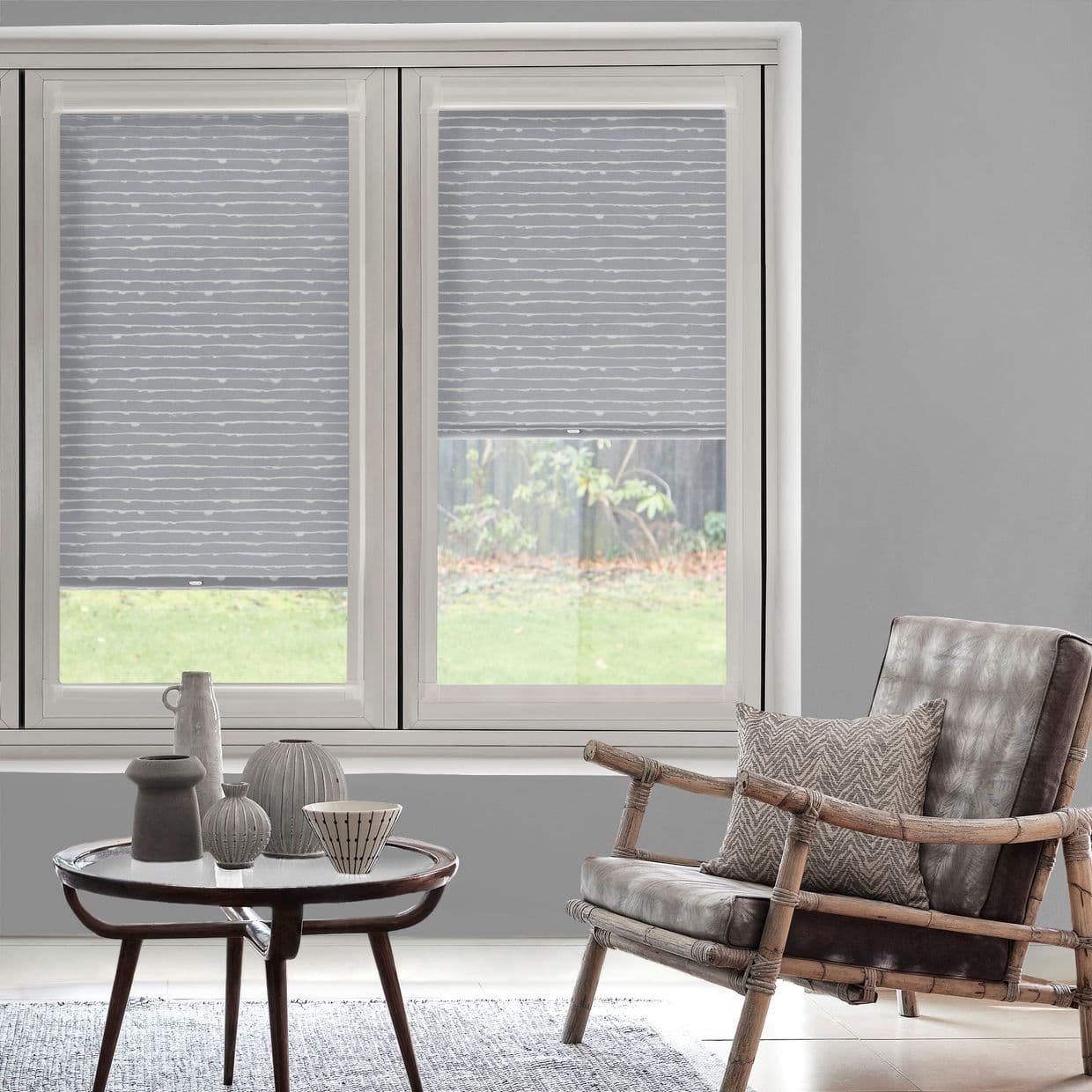 Grey roller blinds with subtle horizontal white lines on dual casement windowsâ€”left fully lowered, right mid-heightâ€”filtering daylight. Softly lit living room with rattan armchair, side table, and backyard lawn visible.