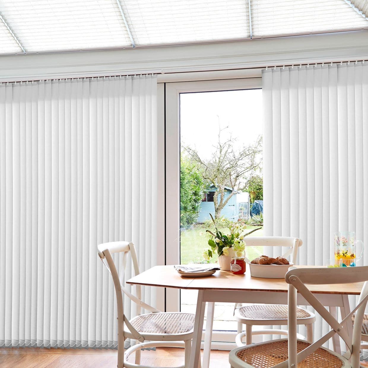 White vertical blinds cover a patio door, mostly closed but parted centrally to reveal outdoors; filtering daylight into a dining area with table, chairs, flowers and garden view.