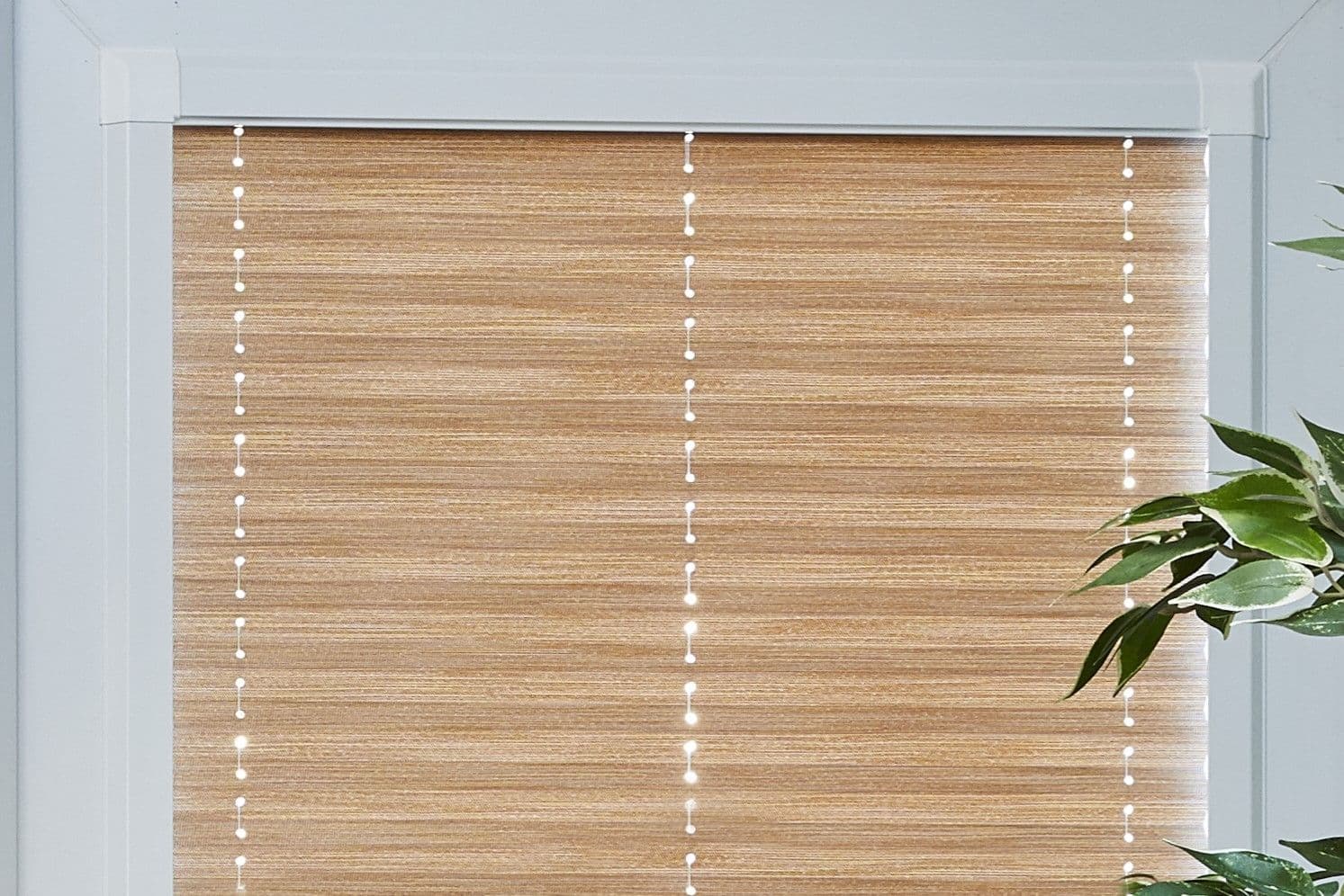 Beige woven roller blind, fully lowered with three vertical bead cords showing tiny light dots, covering a white-framed window; interior room lit by soft daylight, leafy plant at right.
