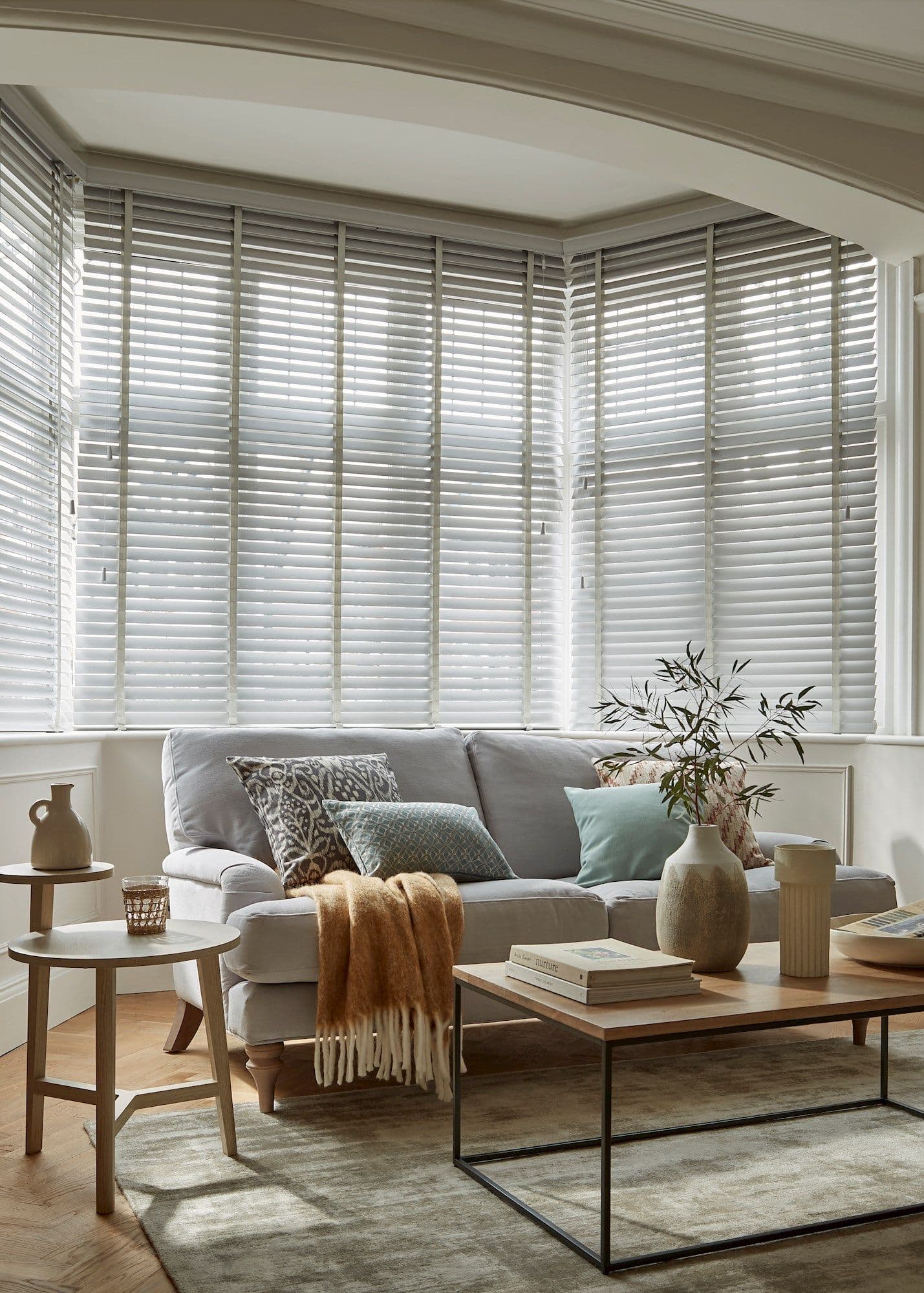 White horizontal Venetian blinds across a bay window, slats mostly closed and filtering soft daylight; in front, a light-gray sofa with cushions, wooden tables, vase, and warm neutral living room.