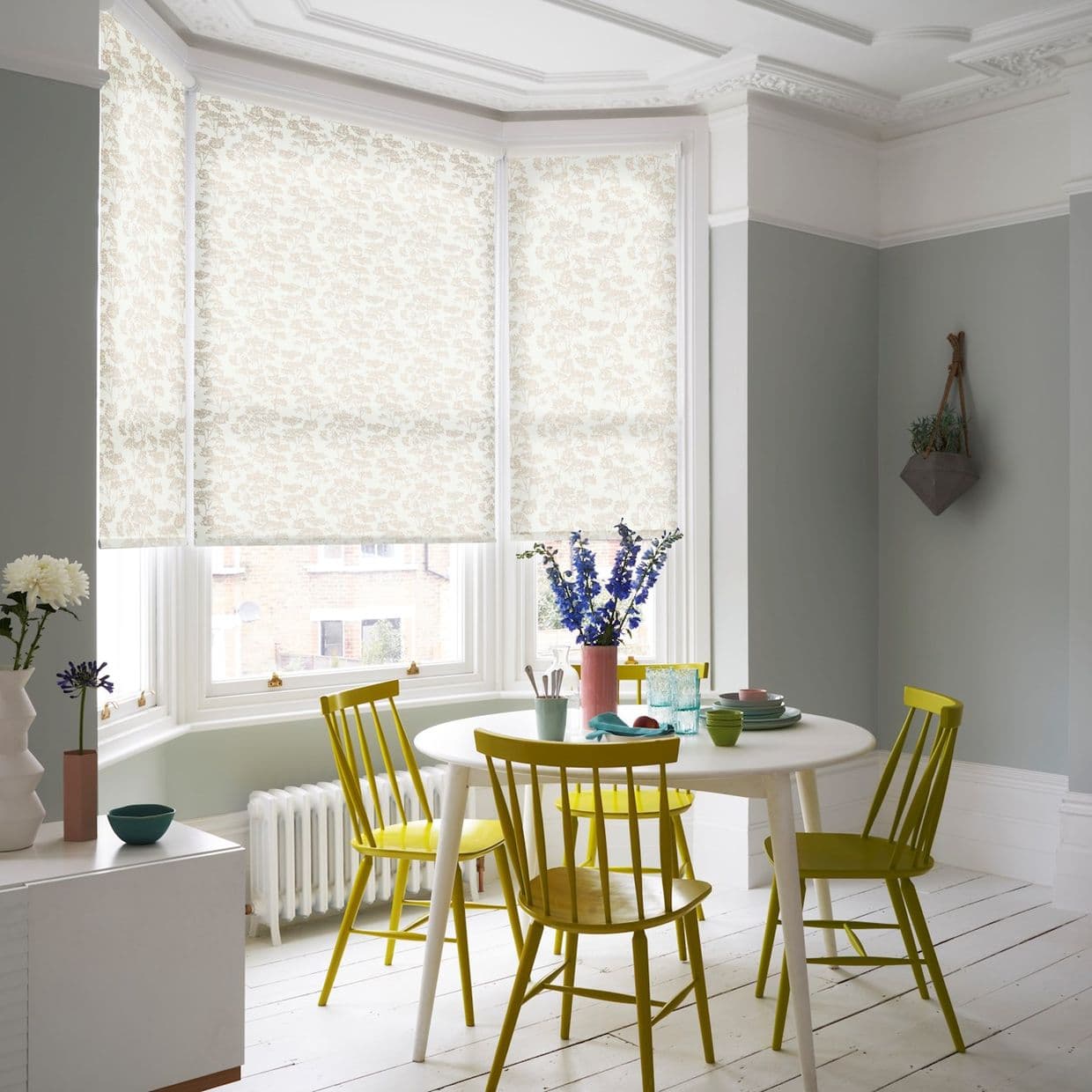 Floral-patterned roller blinds lowered over a three-pane bay window, filtering soft daylight; dining nook with round white table, four yellow chairs, pale grey-green walls and vase of purple flowers.