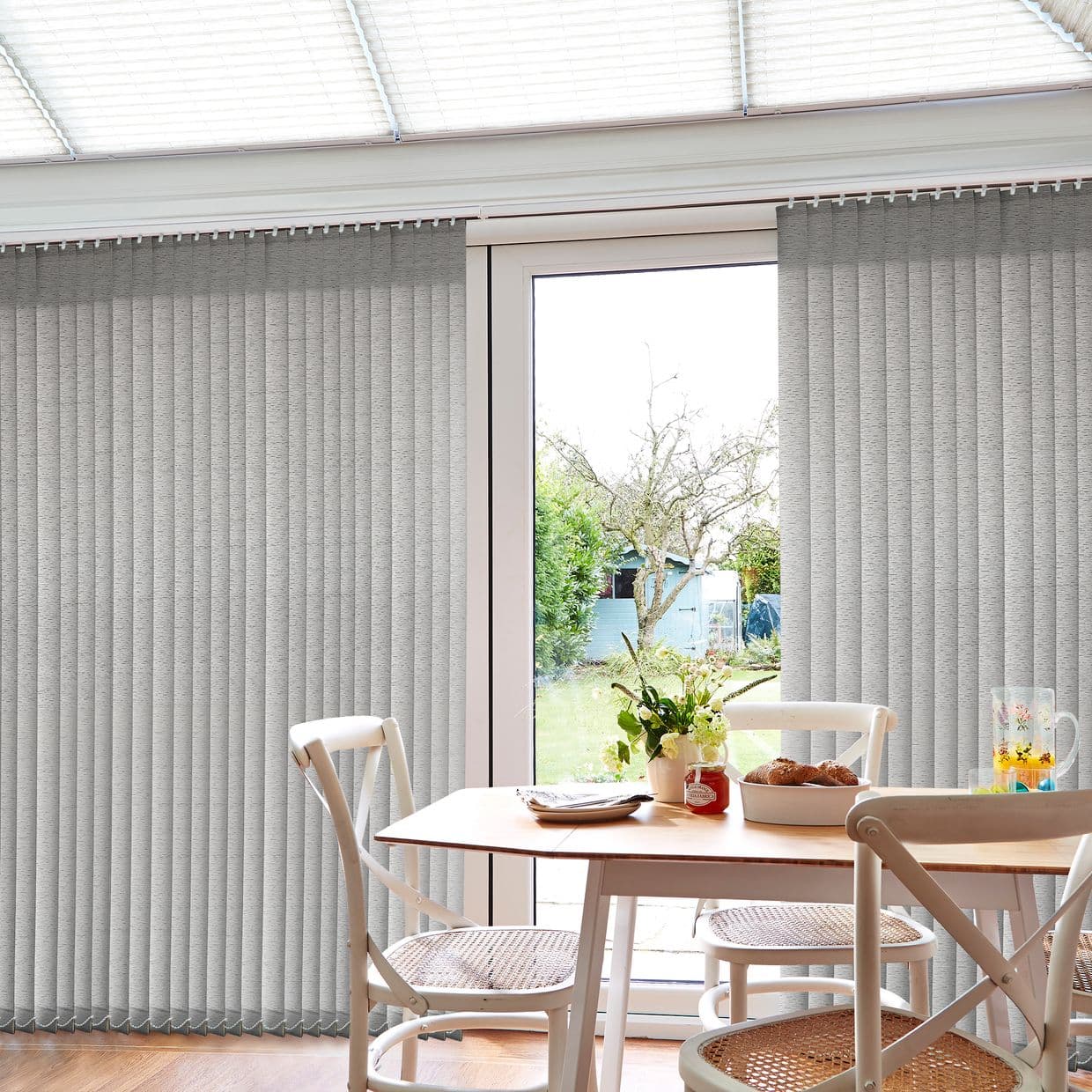 Grey textured vertical blinds cover floor-to-ceiling sliding glass doors, parted centrally to reveal a sunlit backyard; a small wooden dining table with chairs, flowers and pastries sits in the foreground.