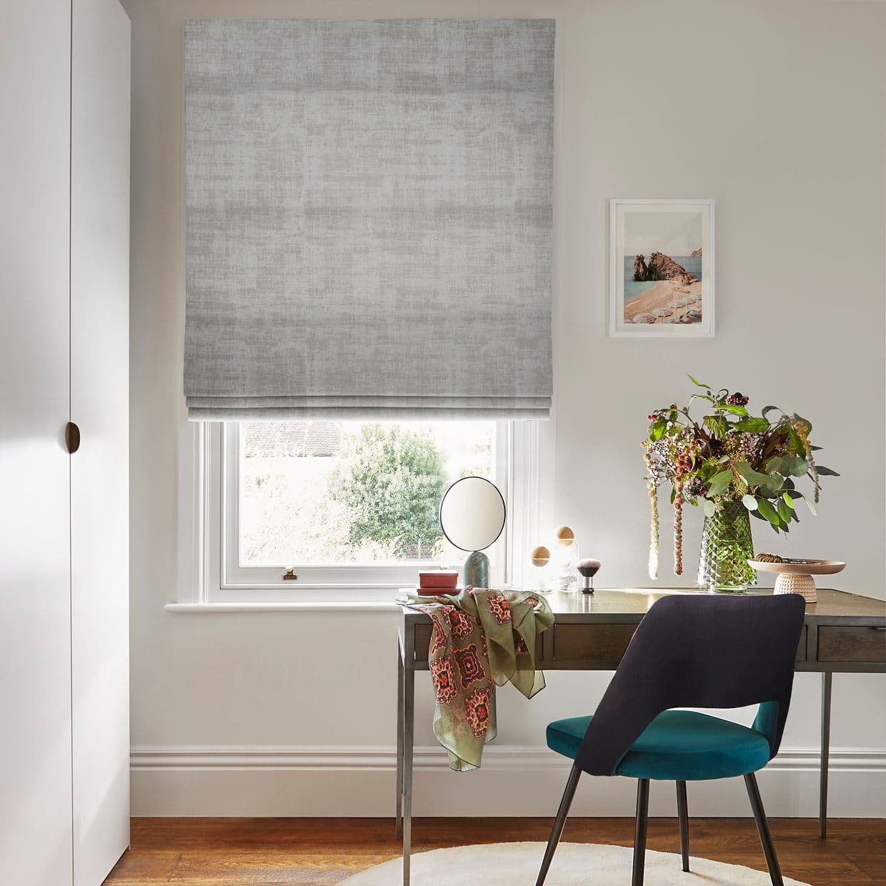 Grey textured Roman blind, lowered in soft folds over the window, filtering daylight; small desk with mirror, teal chair and vase of cascading flowers in a bright neutral home workspace.
