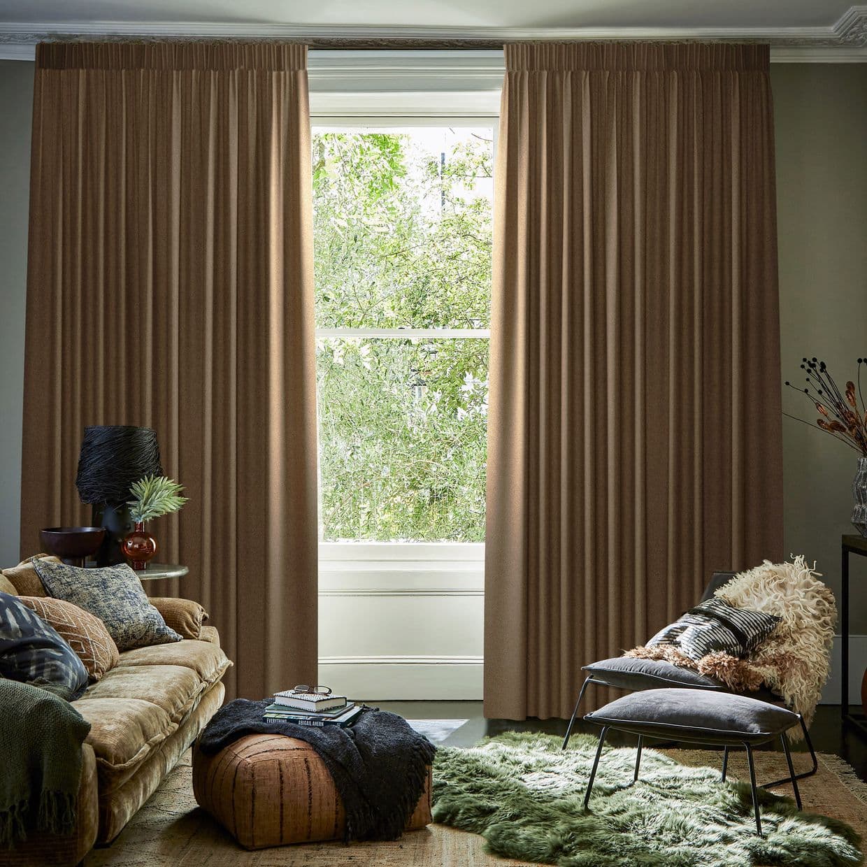 Full-length pleated brown drapes, ceiling-mounted and drawn slightly apart revealing a bright sash window; filtering daylight and a green tree view, in a cozy, earth-toned living room with plush seating.