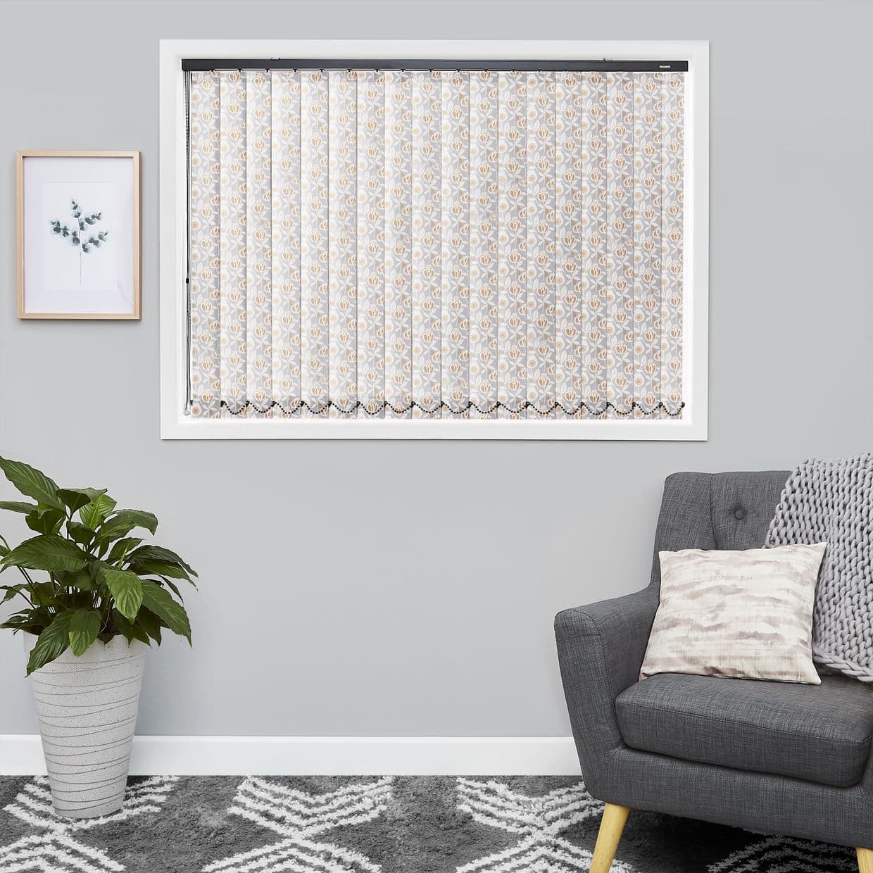 Lace-patterned vertical blinds fully closed across a white-framed window, beige floral motifs filtering soft daylight into a grey living room with armchair, knitted throw, potted plant and framed botanical print.