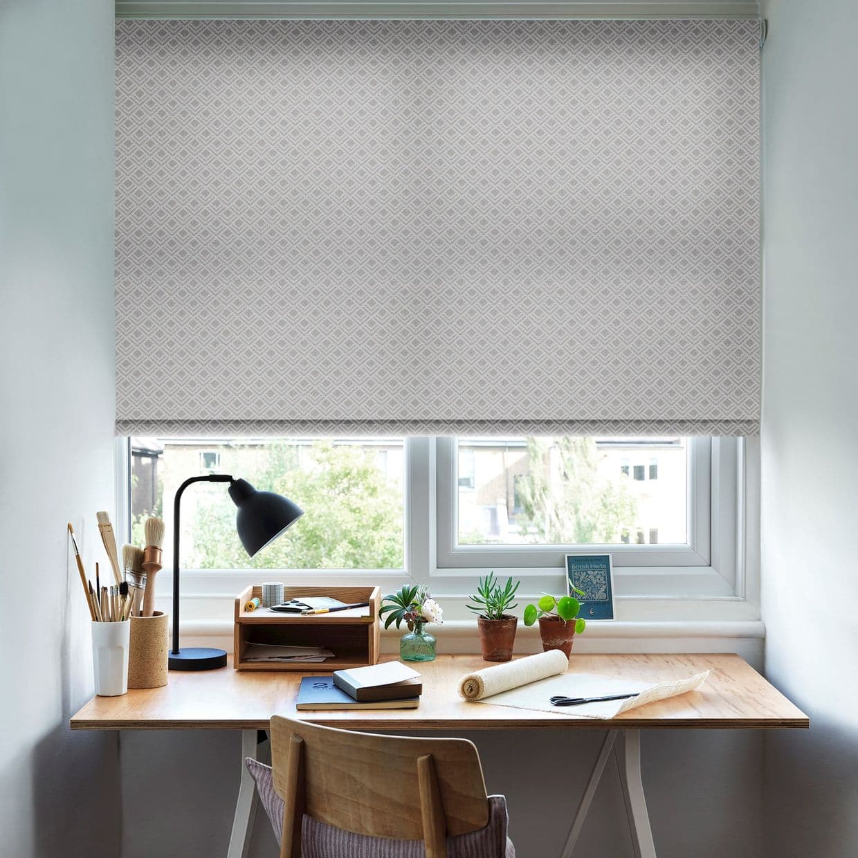 A pale, geometric-patterned roller blind lowered over a two-pane window, filtering daylight; a wooden desk beneath holds a black lamp, paintbrushes, notebooks, small potted succulents, and crafting materials.