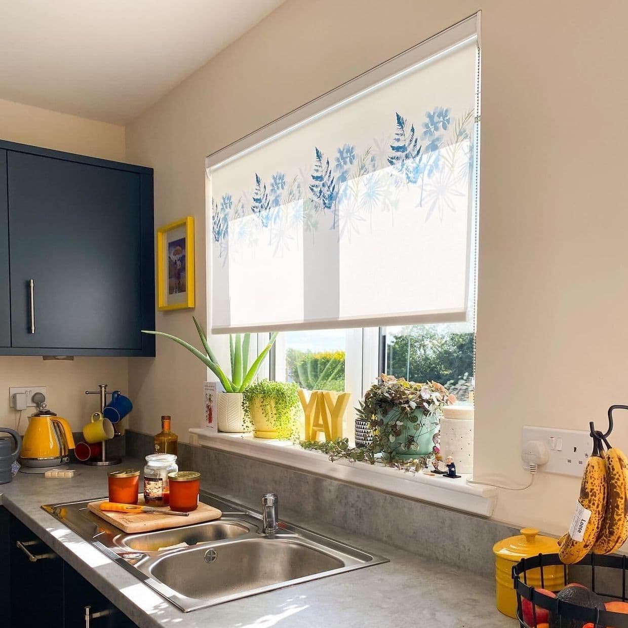 A white roller blind with blue floral and fern motifs, partially lowered across a window, filtering daylight; kitchen sink countertop with potted plants on the sill, garden view beyond. YAY