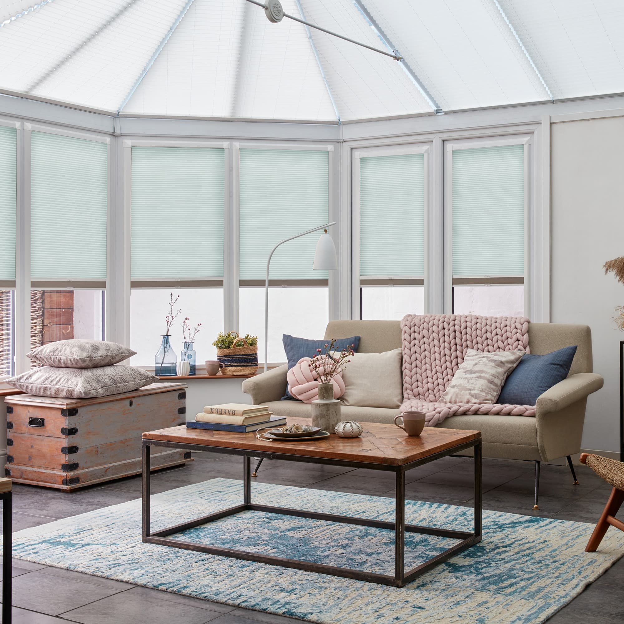 Light-blue cellular blinds cover tall conservatory windows, lowered and filtering soft daylight; a beige sofa, wooden coffee table and pastel cushions occupy a bright, cozy sunroom.