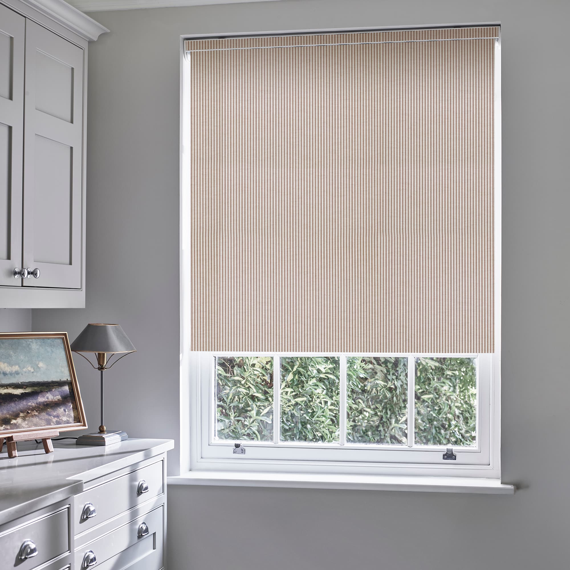 Beige narrow-striped roller blind lowered over most of sash window, leaving lower panes exposed; filters daylight and provides privacy in gray room with white dresser, lamp, and hedge view.