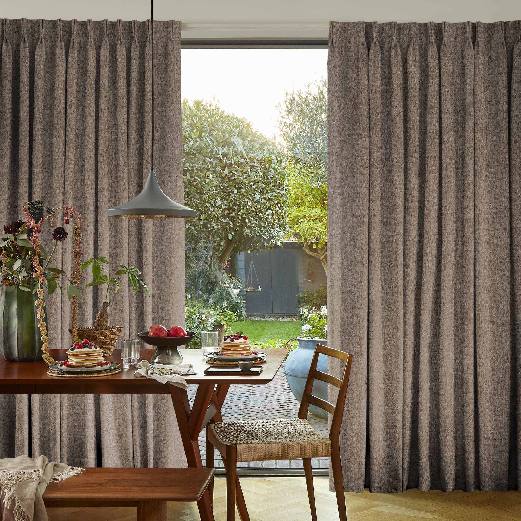 Floor-to-ceiling pleated grey curtains, parted centrally to frame a glass patio door, filtering soft morning light; dining table set with pancakes, fruit and pendant lamp, wooden chair, leafy backyard view.