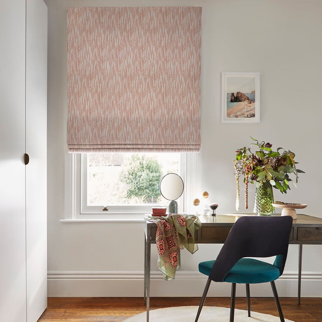 Pink patterned Roman blind, fully lowered and gently filtering daylight over a sash window; wooden desk beneath with mirror, teal chair, vase of foliage, and a framed seaside print on the wall.