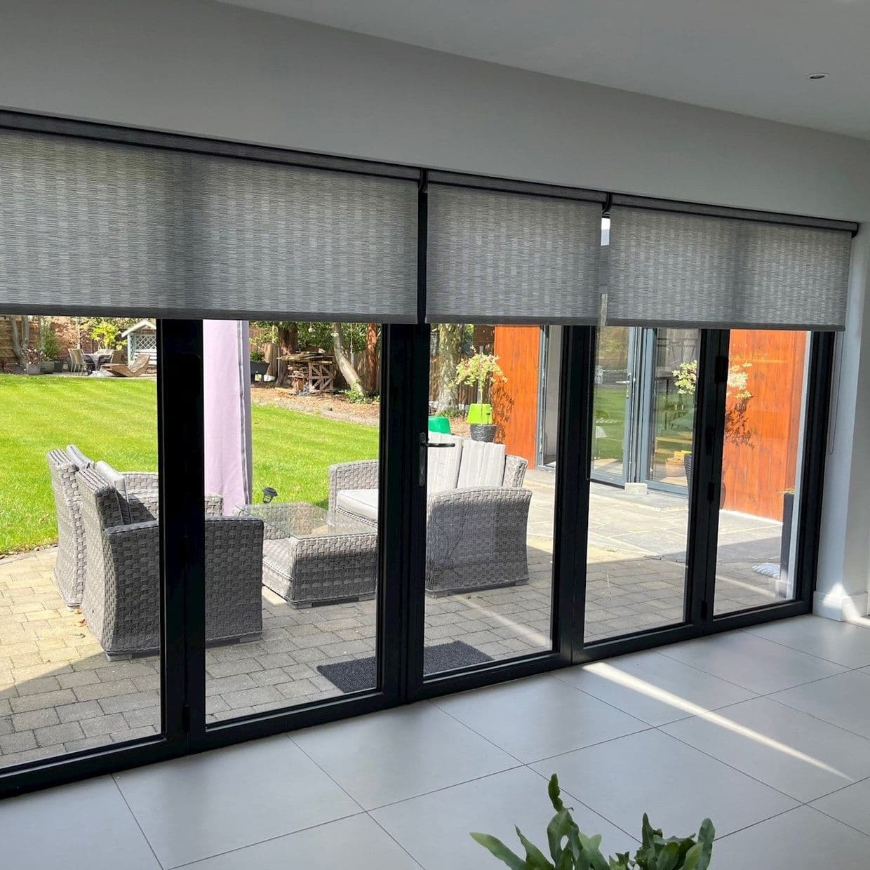 Grey textured roller blinds across four tall sliding glass doors; partially lowered, filtering daylight. Outside, paved patio with wicker seating and green lawn; inside, light-tiled floor and bright modern room.