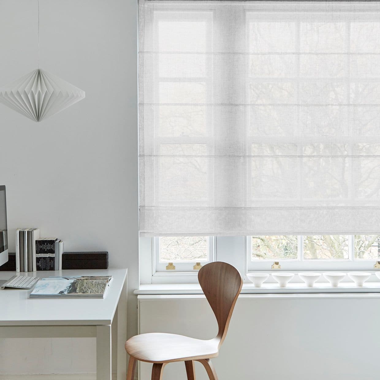 Sheer linen Roman shade, lowered across a multi-pane window, softly filtering daylight through visible tree branches; minimal home office with white desk, wooden chair, and geometric pendant lamp.
