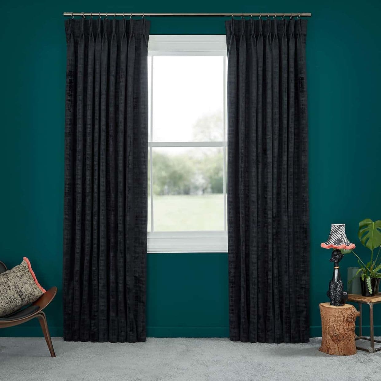 Dark floor-length pleated curtains on a metal rod, drawn to sides and framing a white double-hung window; daytime light through glass, teal-painted wall, carpeted floor, chair and lamp nearby.
