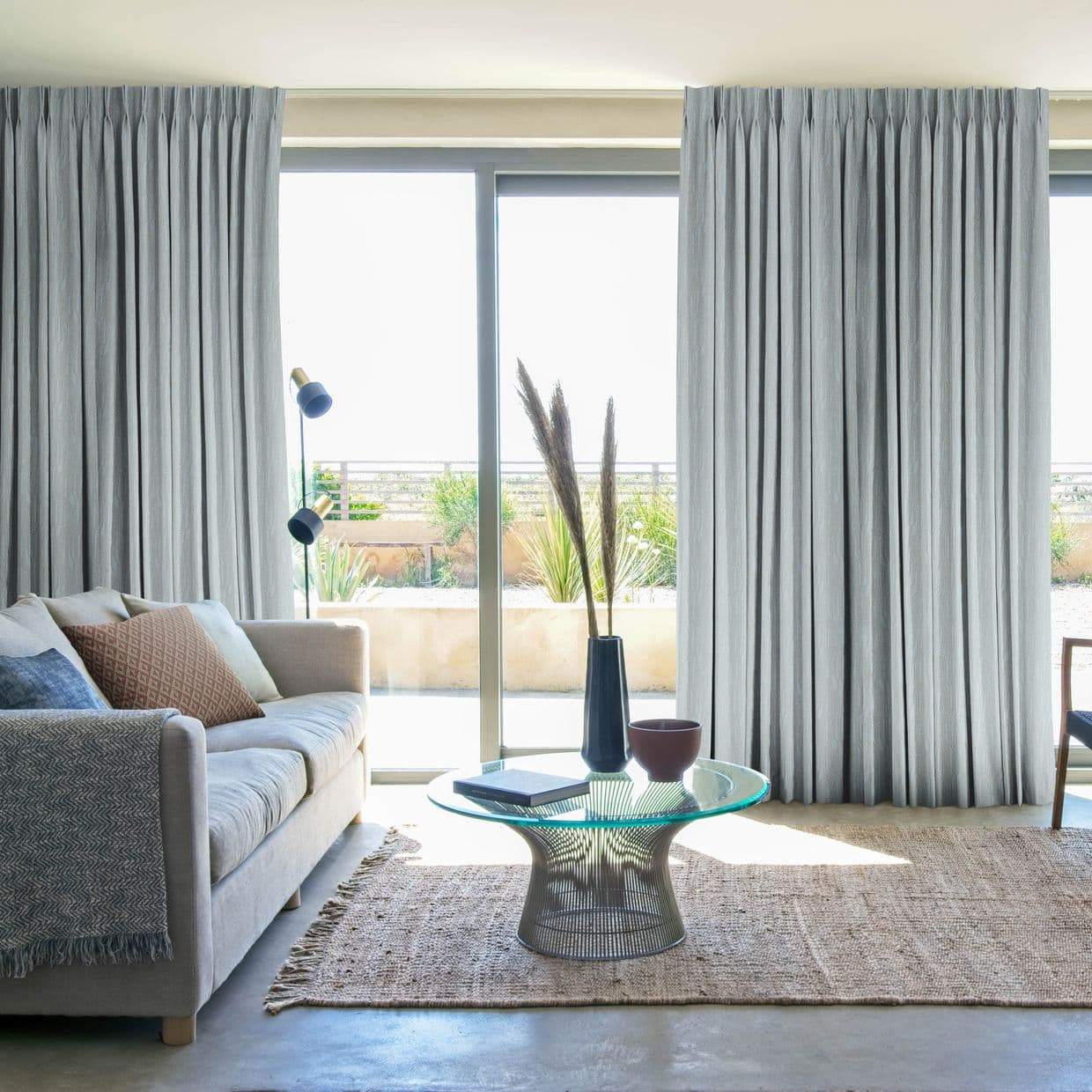 Floor-to-ceiling pleated grey curtains, partly drawn across sliding glass doors, filtering bright daylight into a sunlit living room with sofa, glass coffee table, and a planted outdoor terrace view.