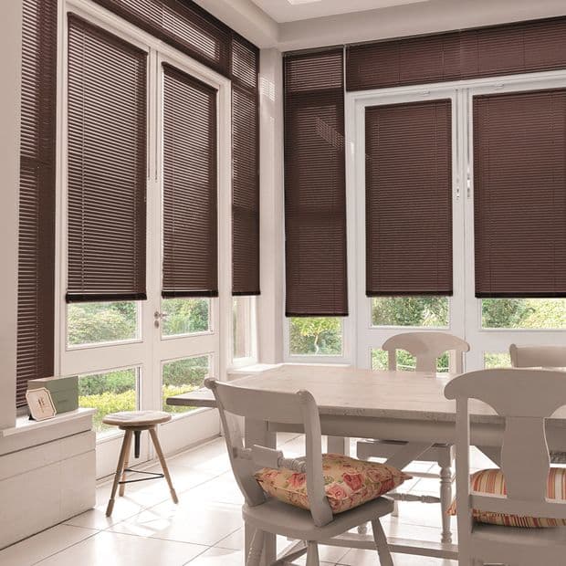 Brown horizontal Venetian blinds on multiple window and door panels, lowered and mostly closed, filtering light; casting shade across a sunlit dining area with table, chairs and garden view.