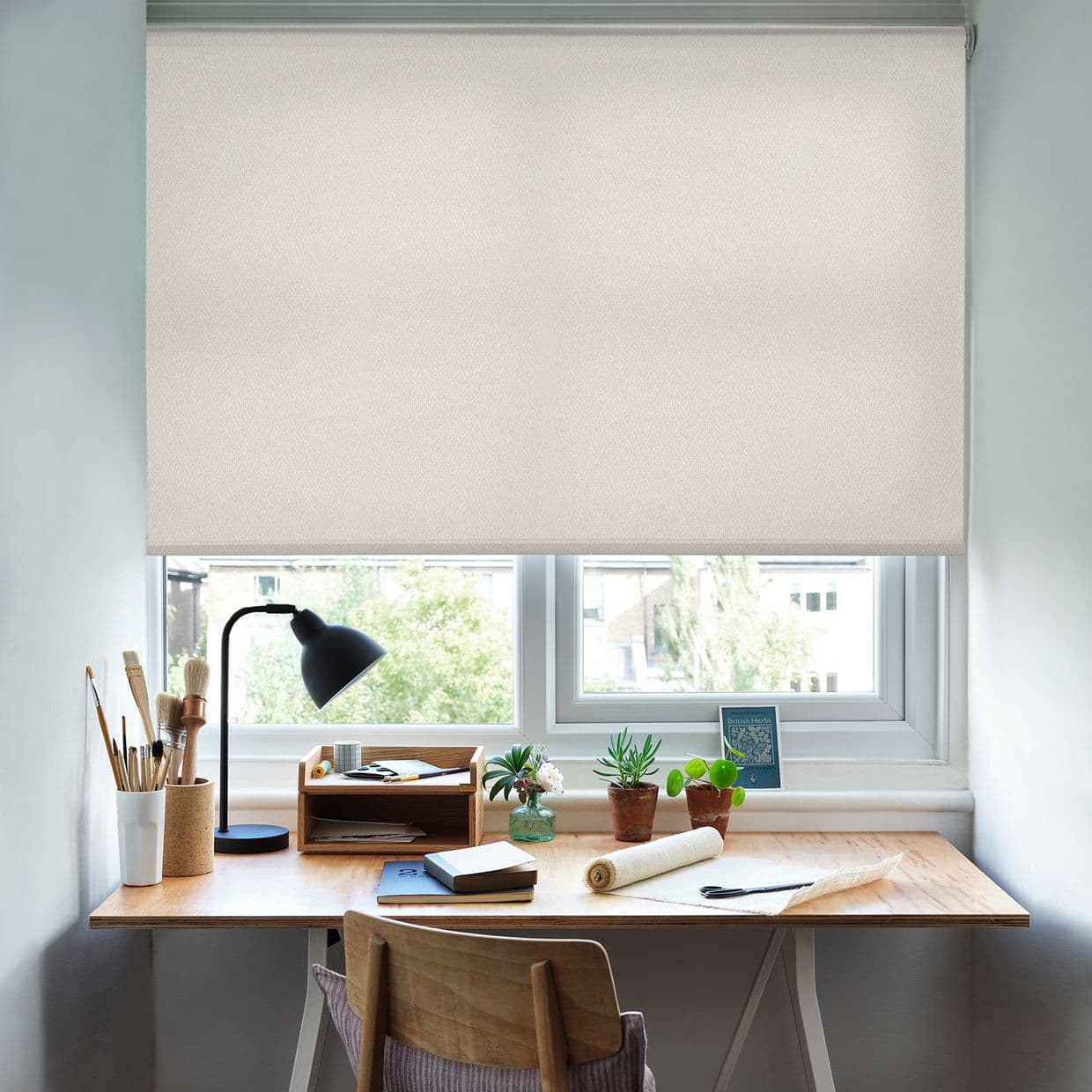 Beige textured roller blind, lowered almost to the sill with a narrow gap; filtering daylight over a tidy wooden desk with lamp, brushes, notebooks, small potted plants, and a chair.