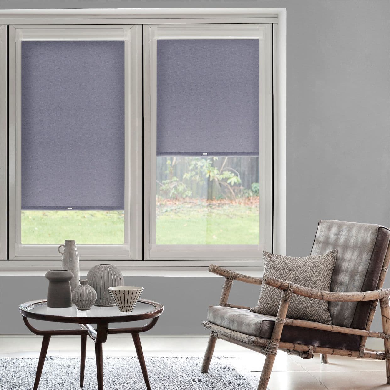 Two fabric roller blinds, lowered mid-height and opaque dusky blue, covering double windows and filtering daylight; cozy living room with rattan armchair, low coffee table, muted gray walls, garden view.