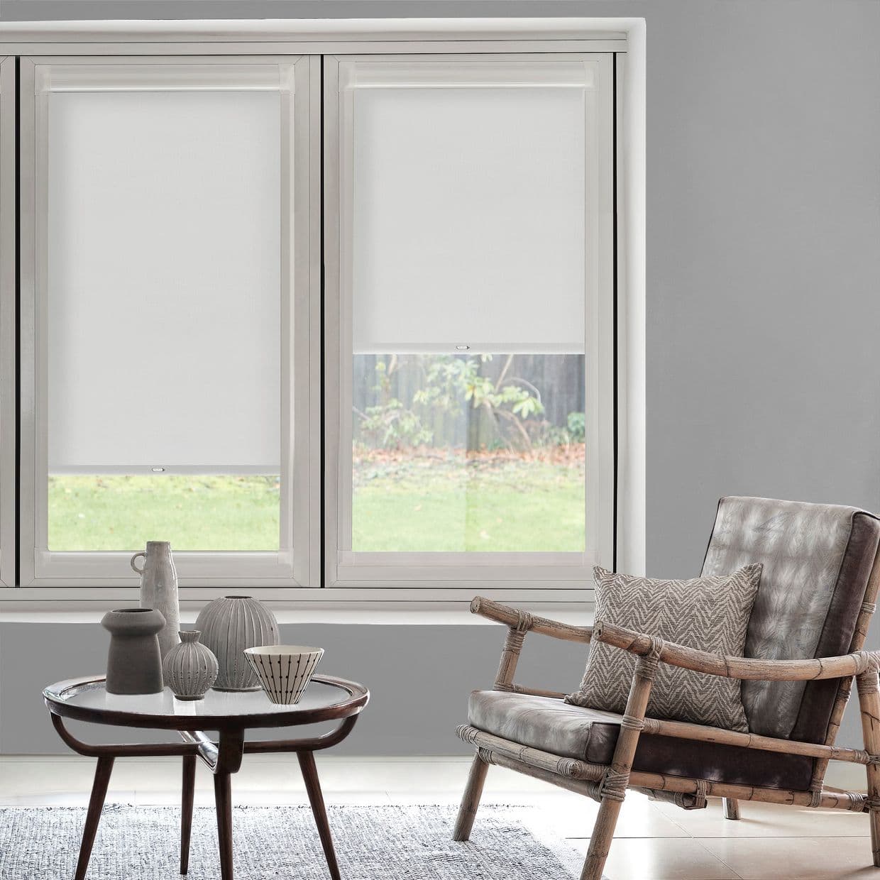 White roller blinds on a twin window, left fully lowered, right halfwayâ€”filtering daylight and obscuring view; modern living room with grey walls, bamboo-framed armchair and small table, grassy yard outside.