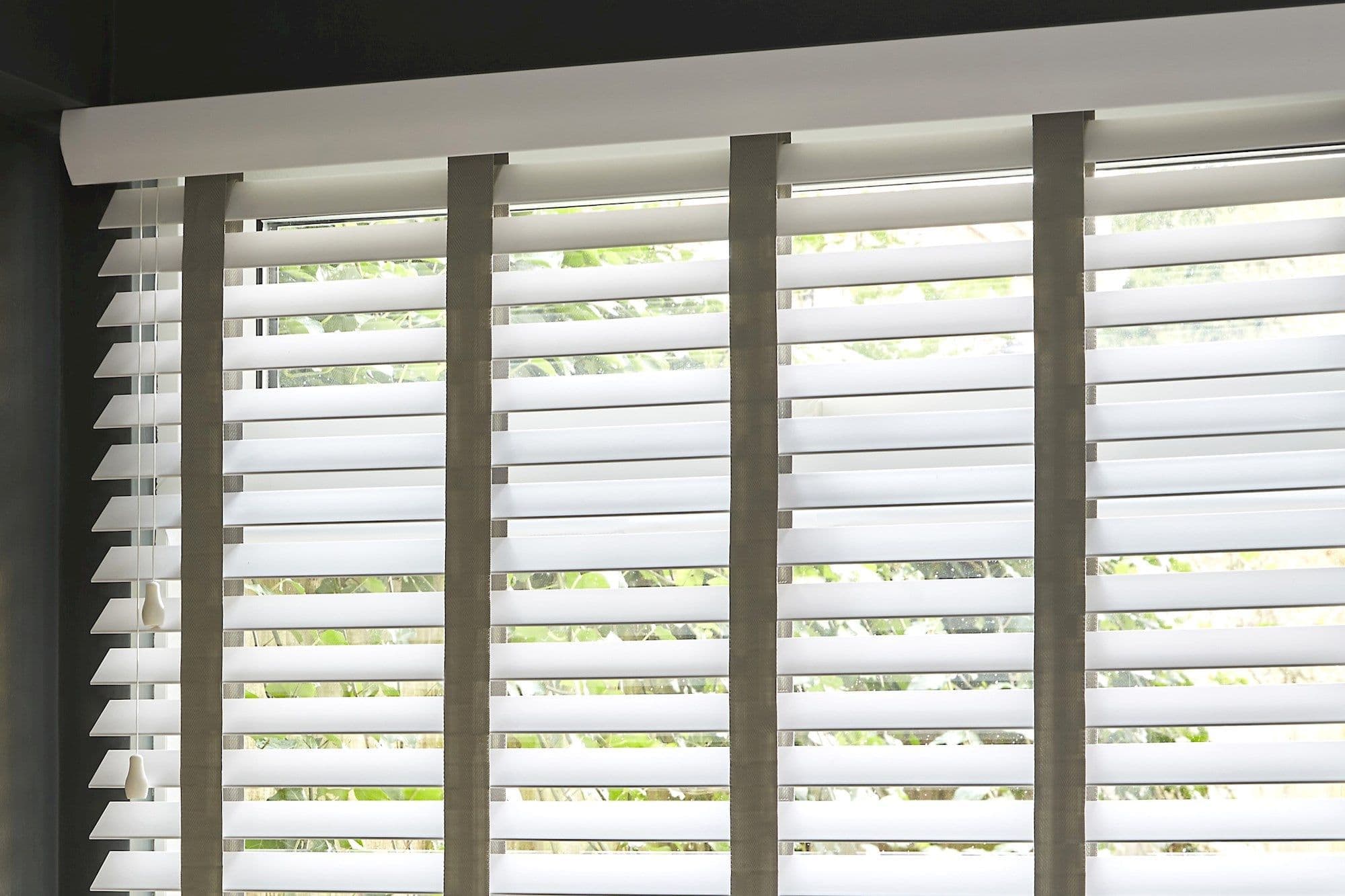 Horizontal white Venetian blinds with wide slats and beige vertical tapes, mounted inside top casing; slats tilted partially open, filtering bright daylight into a room with leafy garden view.