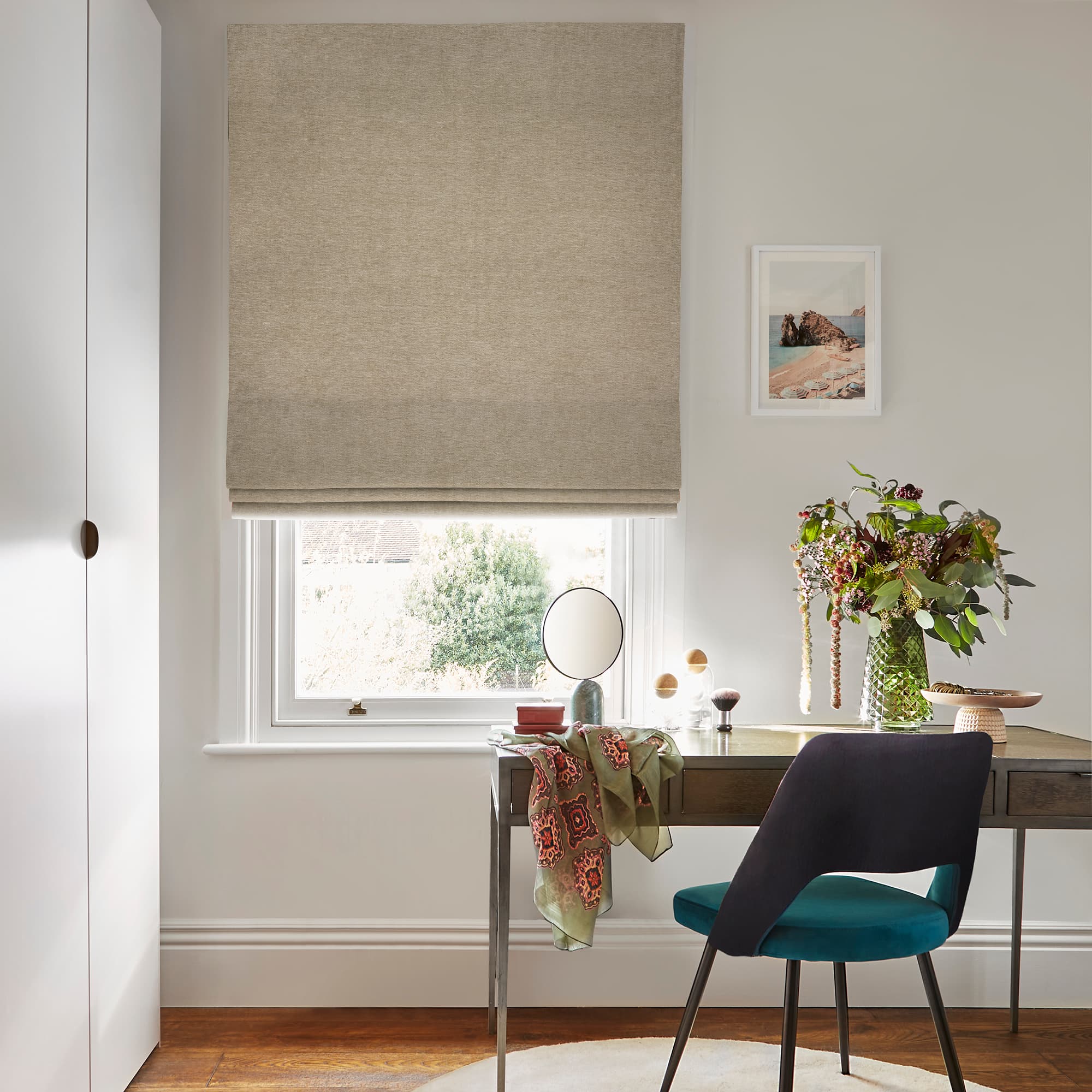 Beige fabric Roman blind lowered and folded, filtering soft daylight through a sash window; desk with teal chair, tabletop mirror and vase of flowers in a calm neutral home office.