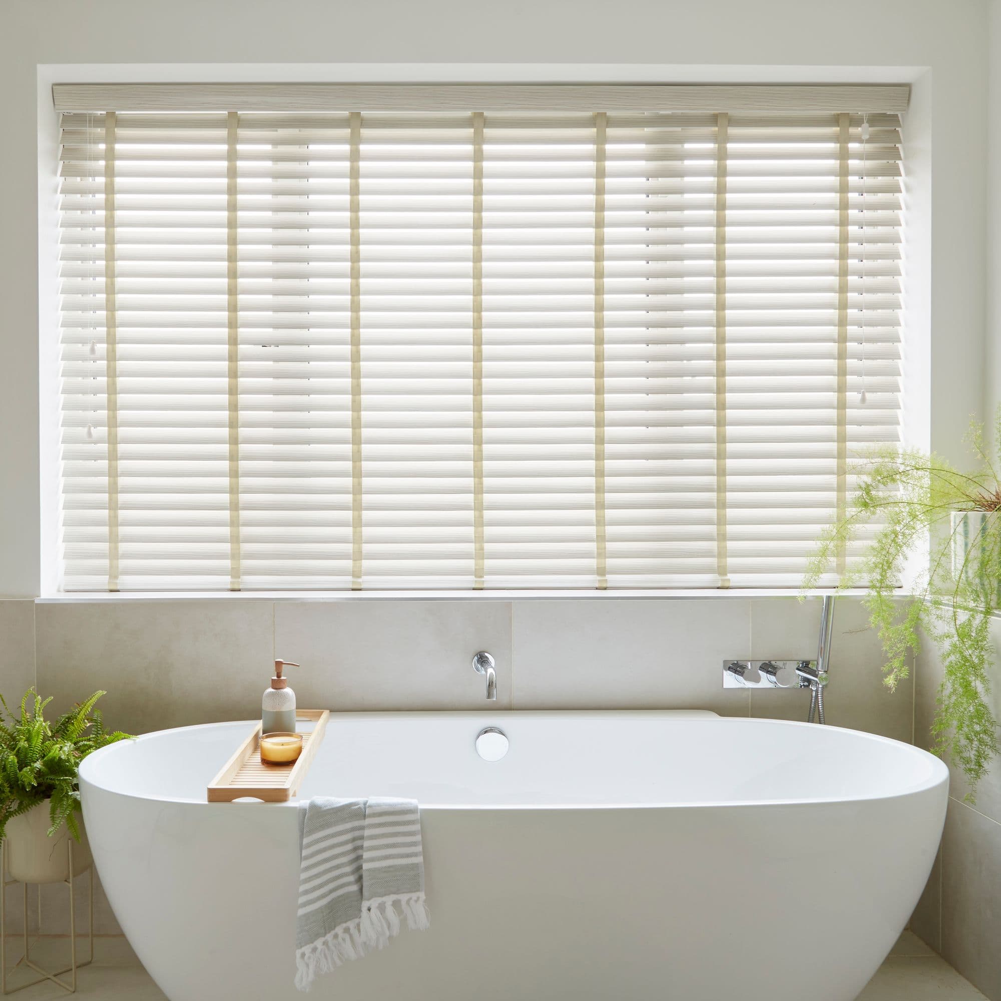 Cream horizontal slatted blinds with vertical ladder tapes cover a recessed window, closed and softly filtering daylight, above a freestanding white bathtub in a neutral, plantâ€‘accented bathroom.