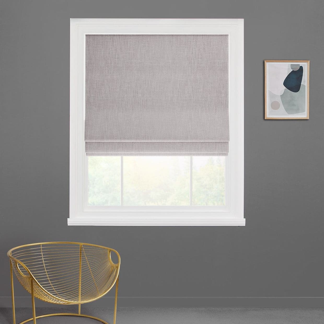 Light-grey fabric Roman shade lowered with a folded bottom, filtering daylight through a white-framed window; on a charcoal wall in a room with a gold wire chair and abstract art.