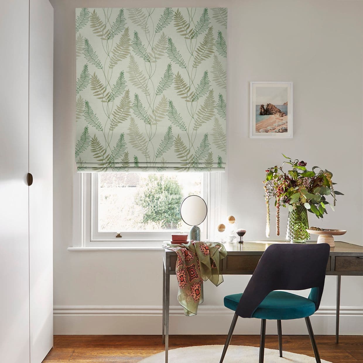 Roman fabric blind with green fern print lowered over a window, filtering daylight; desk with chair, vase of flowers and mirror beneath in a bright home office.