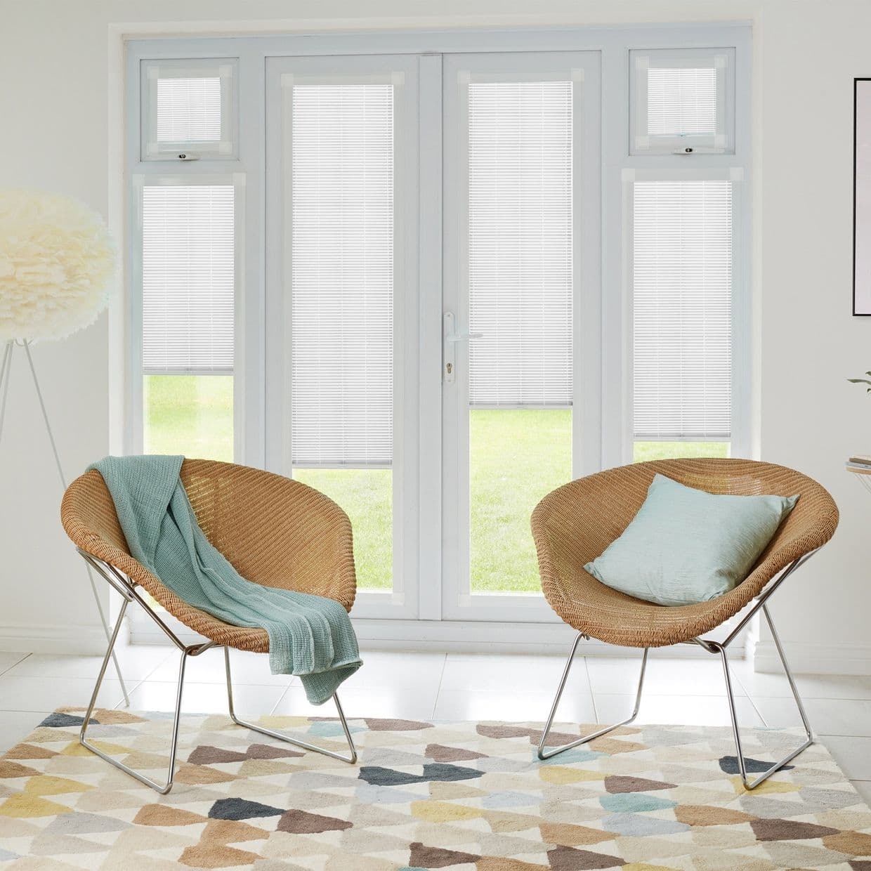 White internal Venetian blinds covering French doors and sidelights, partially lowered and filtering soft daylight; modern bright sitting area with two wicker chairs, pastel cushions, geometric rug and lawn view.